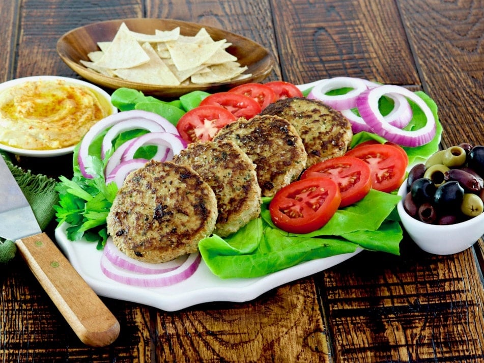 Wide horizontal shot - white plate of four turkey burgers stacked in a line garnished with lettuce, red onion and tomato slices with a dish of olives, hummus, and pita chips on the side, spatula laying nearby on wooden table.