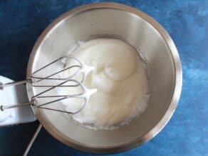 Egg whites whipped to a white frothy texture with electric mixer beaters in a stainless bowl on a blue countertop.