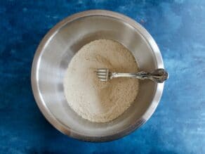 Dry matzo meal and matzo ball ingredients being stirred with a fork in a stainless bowl on a blue countertop.