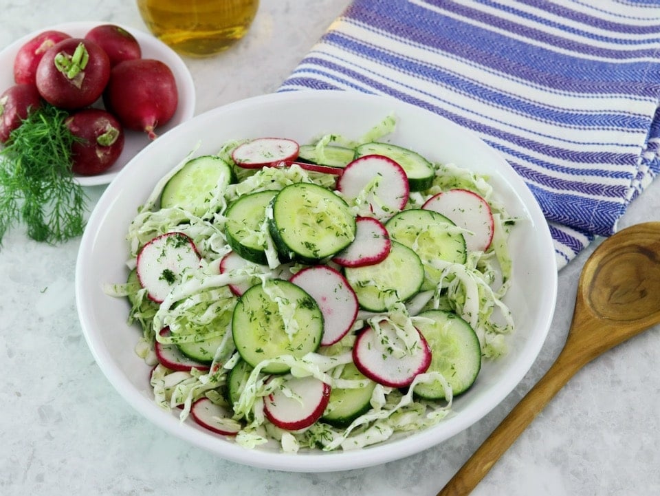 Close up shot of a large bowl filled with crunch pickled salad. A wooden bowl and blue striped napkin sit off to the right side. To the left there is a bunch of fresh radishes.