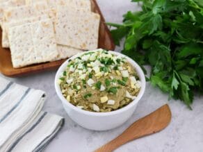 Overhead horizontal shot - White bowl of vegetarian chopped liver topped with chopped hard boiled egg and chopped parsley, with a wooden spatula, bunch of fresh parsley, wooden platter of crackers, and linen napkins beside it, on a marble countertop.