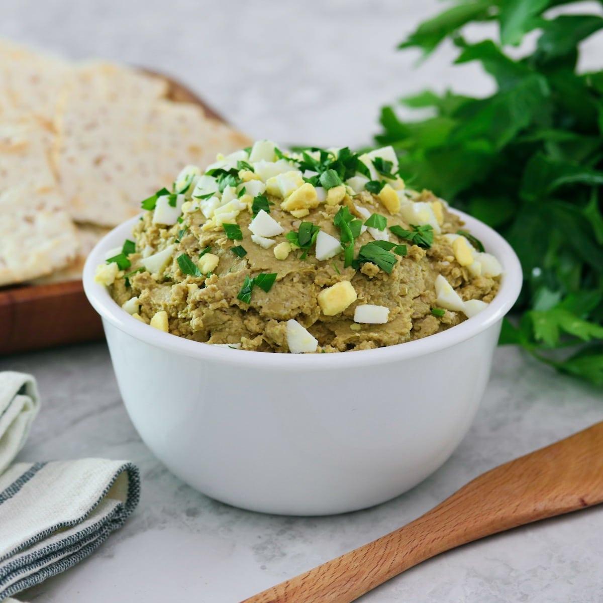 Square featured shot - White bowl of vegetarian chopped liver topped with chopped hard boiled egg and chopped parsley, with a wooden spatula, bunch of fresh parsley, wooden platter of crackers, and linen napkins beside it, on a marble countertop.