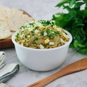 Horizontal shot - White bowl of vegetarian chopped liver topped with chopped hard boiled egg and chopped parsley, with a wooden spatula, bunch of fresh parsley, wooden platter of crackers, and linen napkins beside it, on a marble countertop.