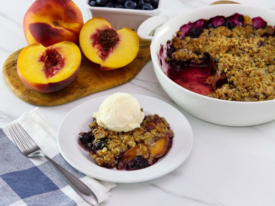 Horizontal image - peach and blueberry crisp on a small white plate, topped with a scoop of vanilla ice cream. A white baking dish of the remaining crisp sits off to the right next to a pile of fresh peaches.