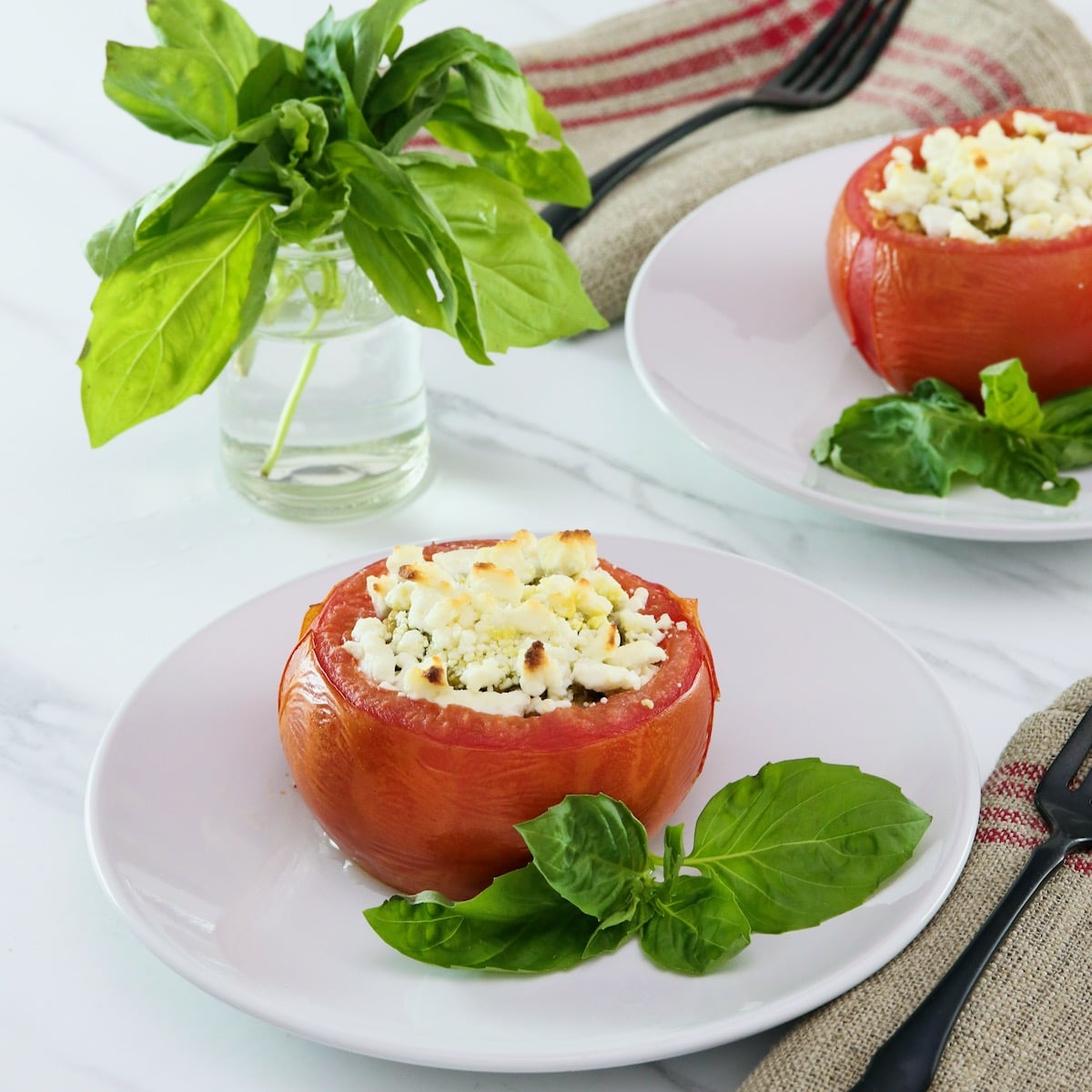 Square image of stuffed tomato on a white plate next to fresh basil leaves. Another plate, a sprig of basil in a glass of water, and a white towel with red stripes in the background.