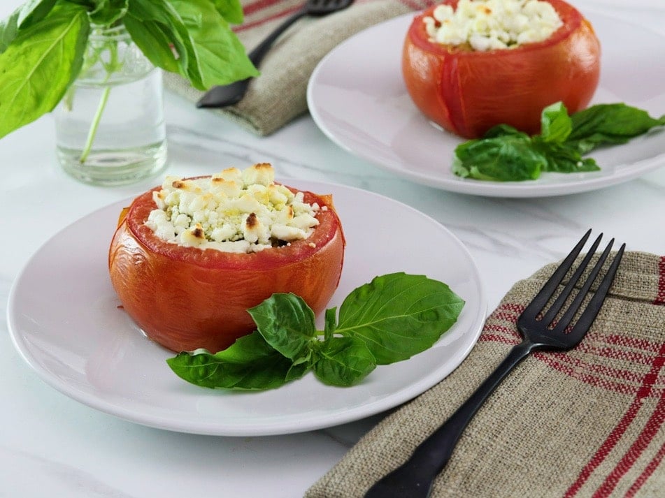 Horizontal image - stuffed tomato on a white plate next to fresh basil leaves, a napkin and fork lay to the right side. Another plate, a sprig of basil in a glass of water, and a white towel with red stripes in the background.