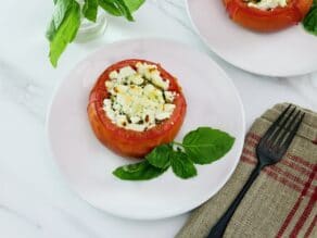 Overhead image - stuffed tomato on a white plate next to fresh basil leaves, a napkin and fork lay to the right side. Another plate and a sprig of basil in the background.