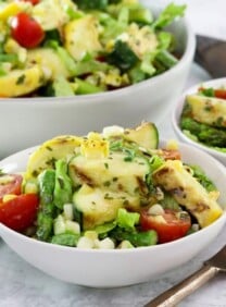 Square image - Two white bowls filled with grilled vegetable salad, a mix of grilled zucchini, squash, asparagus, corn, tomatoes, lettuce and basil with lemony basil dressing. A fork sits off to the right side, a large bowl of the same salad in the background.