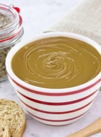 Horizontal shot - bowl of sunflower butter on a marble countertop with wooden spatula, bread slices, and cloth towel. Jar of sunflower seeds in background.