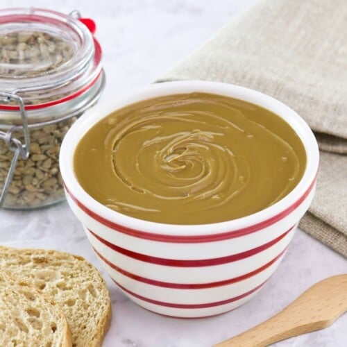 Horizontal shot - bowl of sunflower butter on a marble countertop with wooden spatula, bread slices, and cloth towel. Jar of sunflower seeds in background.