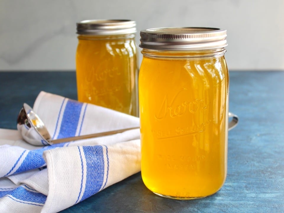 Horizontal shot of two jars of chicken stock on a blue surface with cloth napkin and soup ladling spoon.