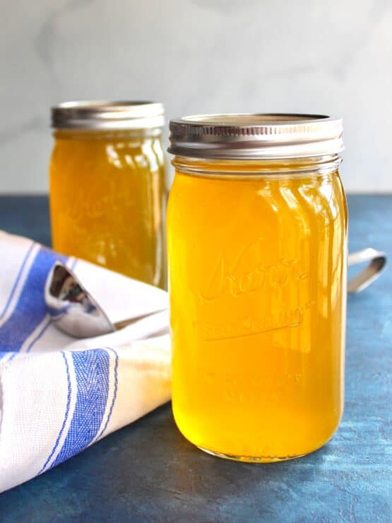 Vertical shot of two jars of chicken stock on a blue surface with cloth napkin and soup ladling spoon.