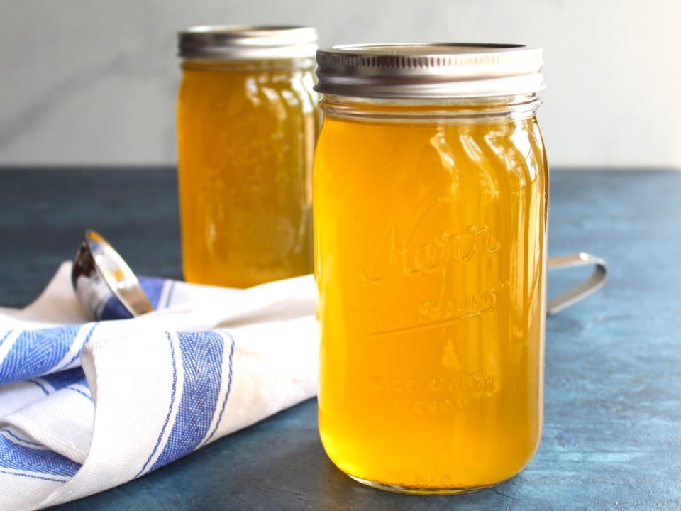 Horizontal shot of two jars of chicken stock on a blue surface with cloth napkin and soup ladling spoon.