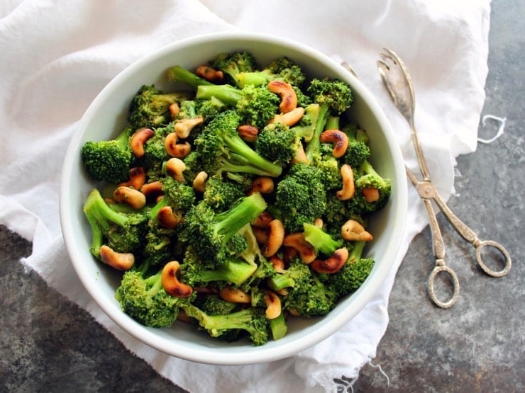 Overhead view of Citrus Broccoli with Cashews in white bowl with antique tongs on grey countertop with white linen. 