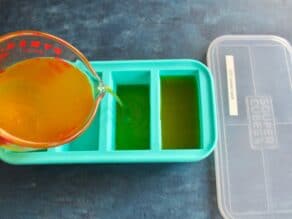 Pouring chicken stock from a measuring cup into a Souper Cubes tray with lid beside, lid labeled with date.