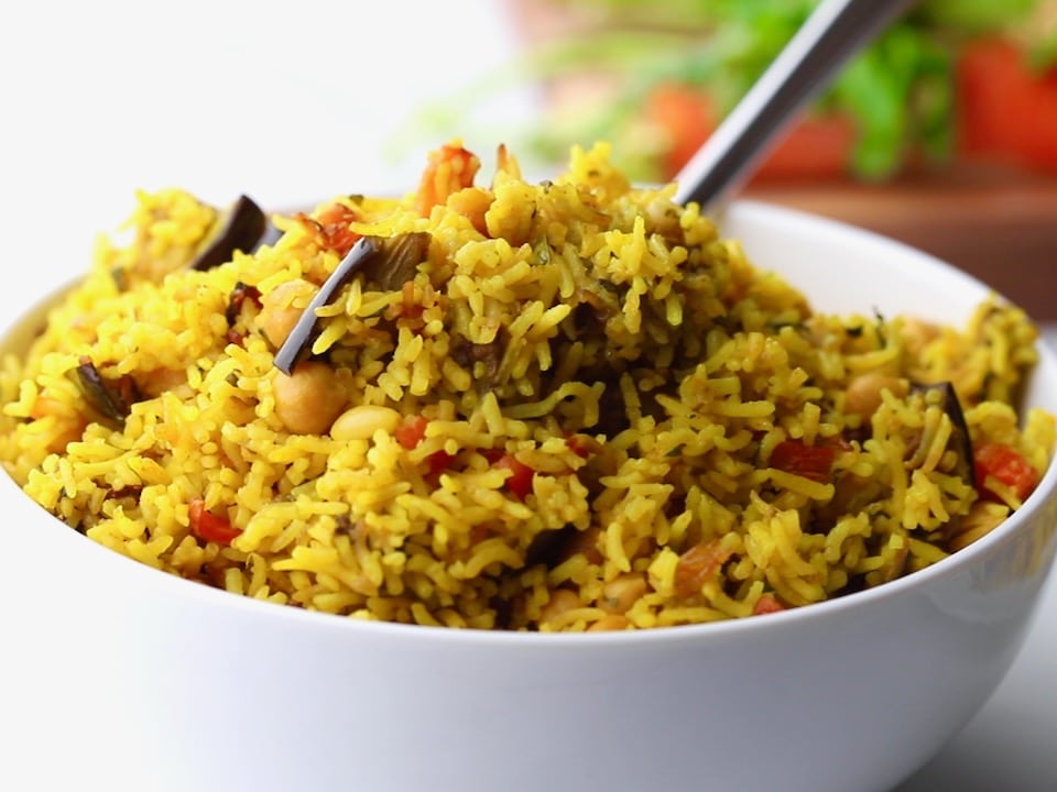 Horizontal close up - spoon digging into fluffy yellow vegetable rice with pieces of eggplant and carrot.