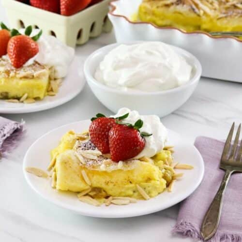 Horizontal shot - a delicious slice of Princess Diana's favorite recipe - bread and butter pudding topped with fresh strawberries and whipped cream. Dish of whipped cream. additional serving, and berries in background.