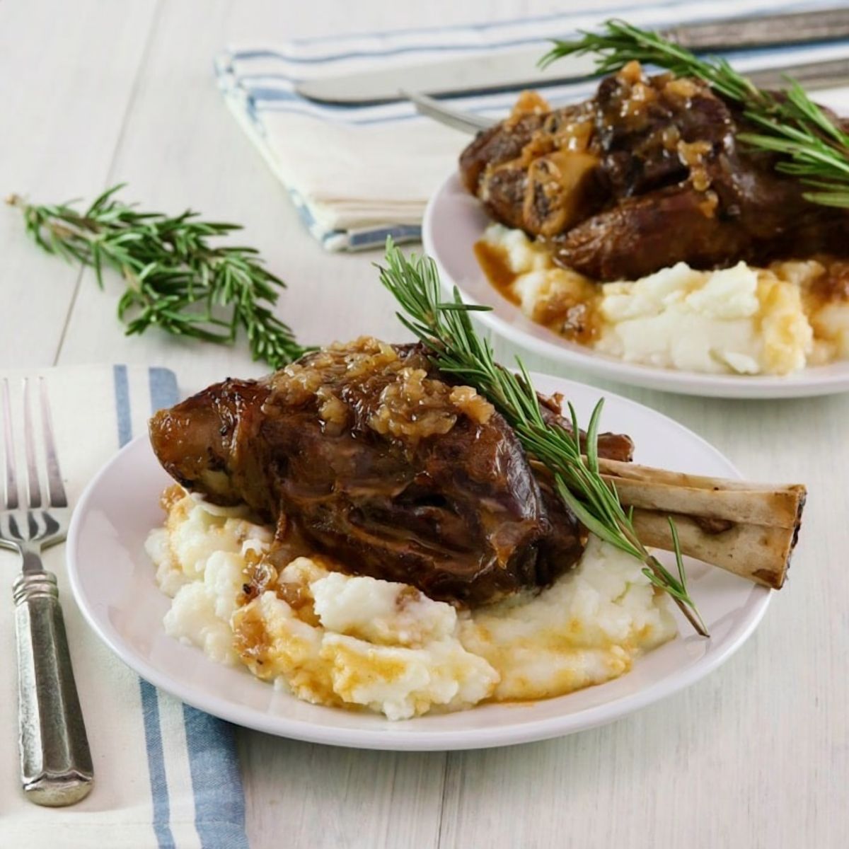 Square featured shot. Tender slow braised lamb shank on mashed potatoes with sprig of rosemary on a white plate, blue and white cloth napkin and fork on white table beneath. Another plate with shank and a sprig of rosemary in background.