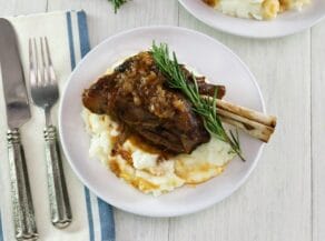 Overhead shot. Braised lamb shank on mashed potatoes with sprig of rosemary on a white plate, blue and white cloth napkin and fork on white table beneath.