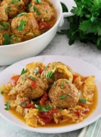 Square image of a white bowl filled with "unstuffed cabbage". A bunch of fresh parsley sits in the back left corner, a larger serving dish of unstuffed cabbage sits in the back left corner..