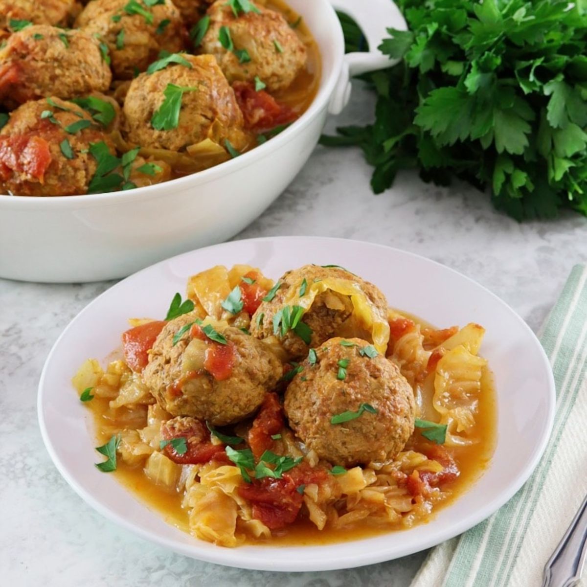 Square image of a white bowl filled with "unstuffed cabbage". A bunch of fresh parsley sits in the back left corner, a larger serving dish of unstuffed cabbage sits in the back left corner..