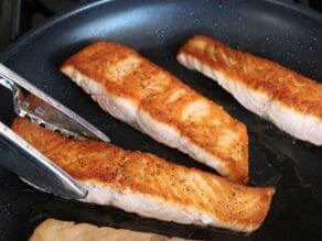 Tongs turning salmon fillet in skillet of seared crispy golden fillets.