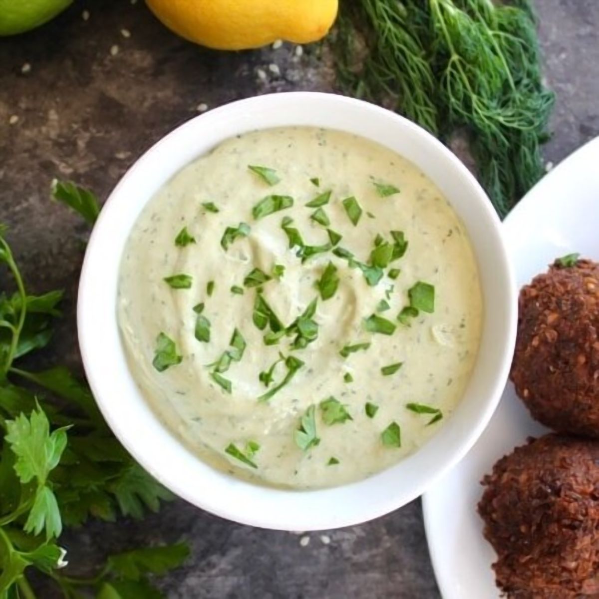 White bowl of herb tahini sauce topped with chopped fresh parsley alongside a falafel plate, fresh parsley, fresh dill, lemon and lime on the side.