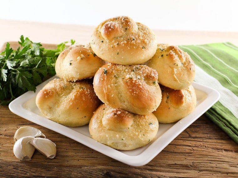 Stocked Garlic Knots topped with cheese and parsley served on a white plate surrounded with parsley and garlic cloves