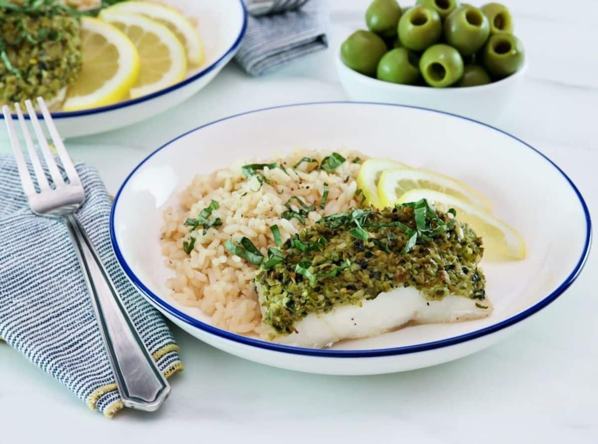 White fish fillet topped with basil tapenade in a bowl alongside couscous and lemon slices.