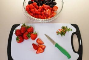 Overhead shot of sliced and hulled strawberries and a green paring knife sitting on top of a cutting board next to a bowl of berries.