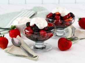 Horizontal shot of two glass dessert dishes filled with a mixture of berries and topped with whipped cream. Two whole strawberries, a dish of whipped cream, and a green tea towel are in the background.