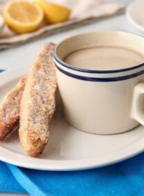 Horizontal shot of a white plate holding a cup of tea and two pieces of mandel bread sitting on top of a blue cloth napkin.