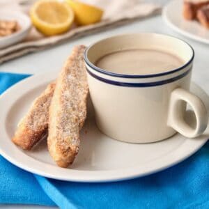 Horizontal shot of a white plate holding a cup of tea and two pieces of mandel bread sitting on top of a blue cloth napkin.