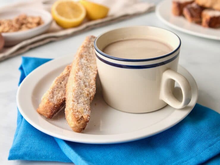 Horizontal shot of a white plate holding a cup of tea and two pieces of mandel bread sitting on top of a blue cloth napkin.