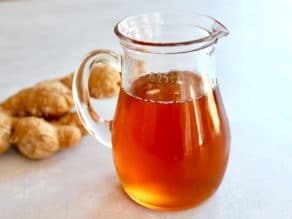 Glass carafe of ginger agave syrup on concrete background with whole ginger root behind.