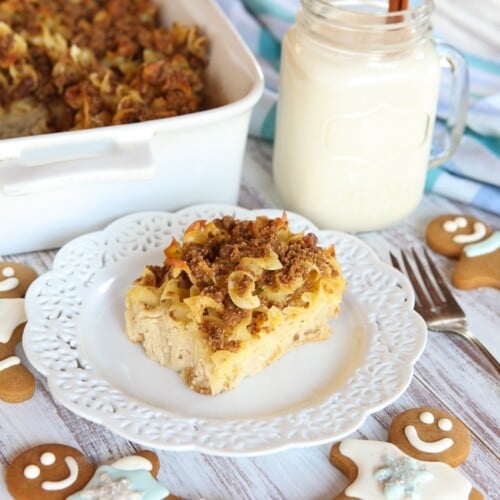 A slice of Eggnog Gingerbread Kugel served on a fancy white plate with fork in the side, a cup of milk, and surrounded with ginger cookies