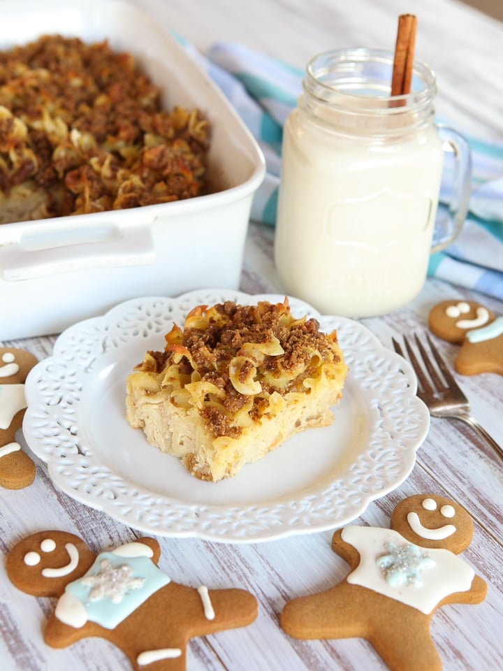 A slice of Eggnog Gingerbread Kugel served on a fancy white plate with fork in the side, a cup of milk, and surrounded with ginger cookies