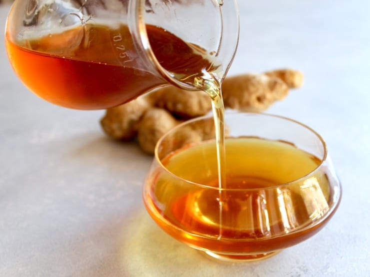 Horizontal shot of glass carafe pouring agave ginger syrup into bowl, whole ginger root in background.