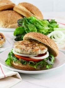 Horizontal shot of a turkey burger on a bun with lettuce, tomato and onion. In the background there is a plate of buns, a plate of sliced tomato and onion, and a plate of turkey burger patties.