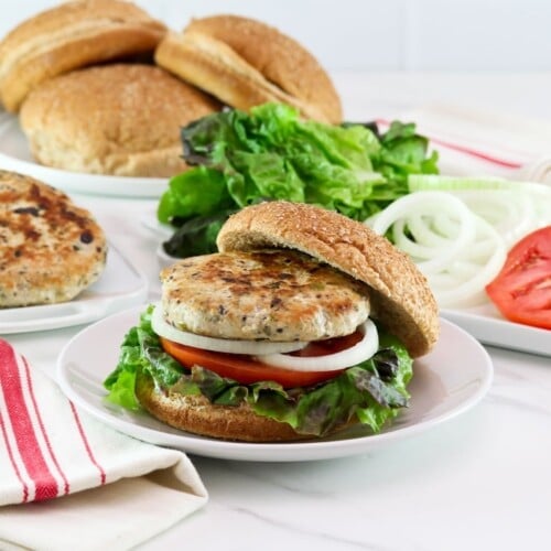 Horizontal shot of a turkey burger on a bun with lettuce, tomato and onion. In the background there is a plate of buns, a plate of sliced tomato and onion, and a plate of turkey burger patties.