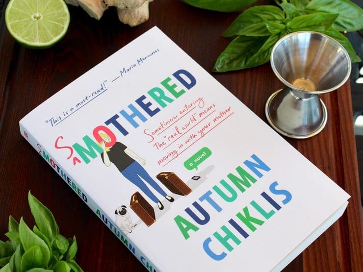 Book - Smothered by Autumn Chills on wooden cutting board with fresh basil, metal jigger, half lime and whole ginger root.