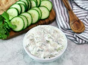 Horizontal shot - white bowl of cucumber raita with cucumber slices in the background.