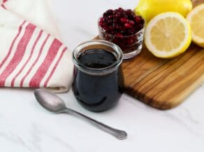 Horizontal shot of a small glass jar filled with pomegranate molasses sitting next to a cutting board holding sliced lemons and a dish of pomegranate arils.