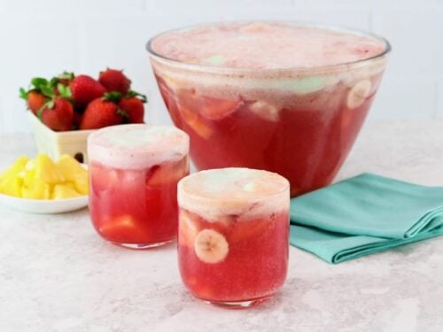 Horizontal shot of a punch bowl filled with a red foamy beverage. Two glasses of the punch sit next to the bowl next to a dish of fresh berries.