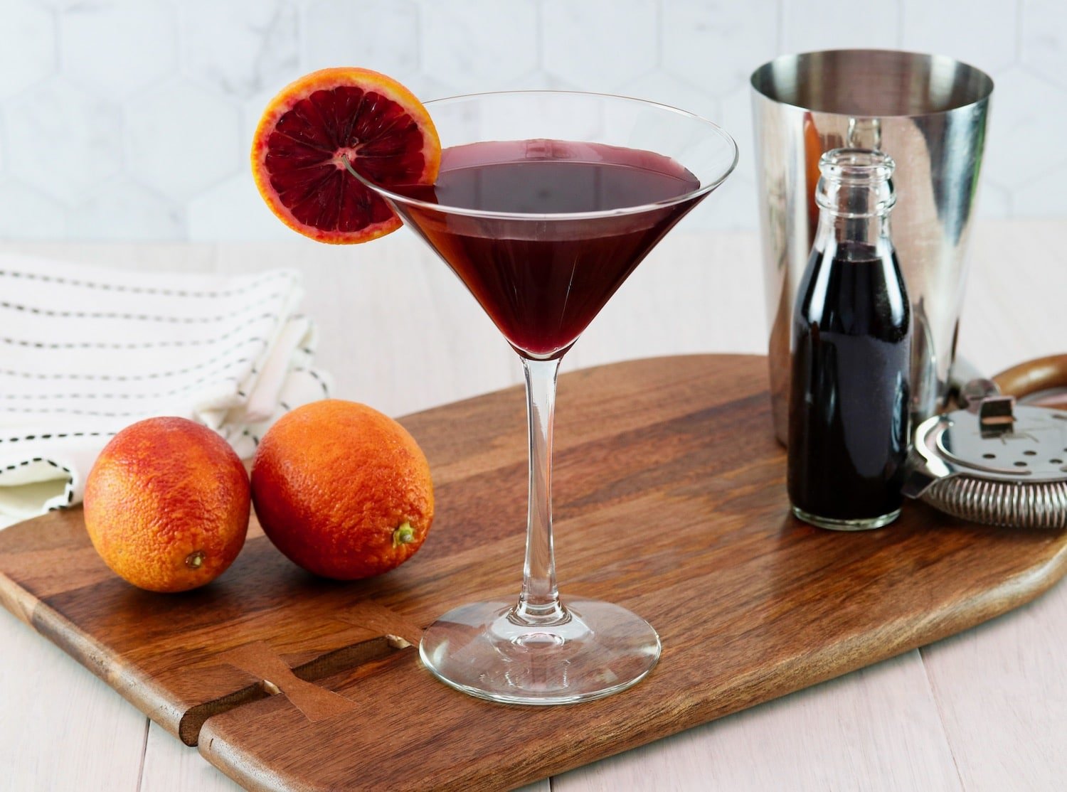 Horizontal image of a martini glass filled with a dark red cocktail and garnished with a slice of blood orange. Two blood oranges and a steel cocktail jigger sit off to the sides.