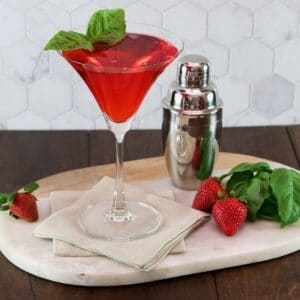 Horizontal shot of a strawberry basil cocktail in martini glass, garnished with a sprig of basil. A steel cocktail shaker sits off to the right side.