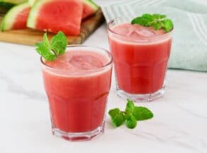 Horizontal shot of two short glasses filled with a frozen watermelon rum cocktail, garnished with fresh mint. Slices of watermelon sit on top of a wooden cutting board on the left side.