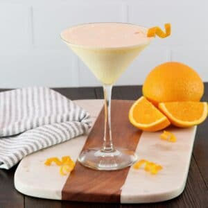 Horizontal shot of an orange creamsicle cocktail in a martini glass garnished with orange twist, on a wood and marble cutting board. Towel and orange with slices in background.