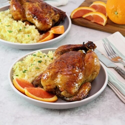 Horizontal shot of a plate containing a citrus marinated game hen on a bed of rice, garnished with fresh orange slices. Another plate of the same food sits in the background next to orange slices on a wooden cutting board.