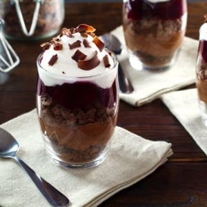 Dark chocolate cherry cheesecake parfait in a glass dessert dish on top of a white napkin next to a spoon on a wooden table.